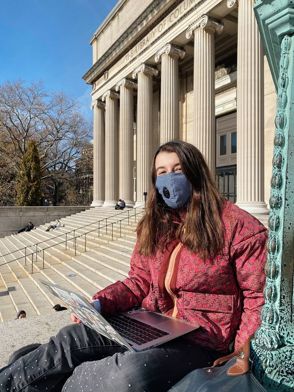 Rachel Barkin sitting on the steps of the Low Library at Columbia with her laptop wearing a puffy red floral jacket, jeans, and a face mask.